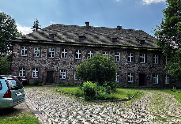 Das große Gebäude aus rotem Naturstein steht im Sonnenschein, umgeben von Bäumen, einer Grünfläche und einem kleinen runden Kiesweg im Vordergrund.