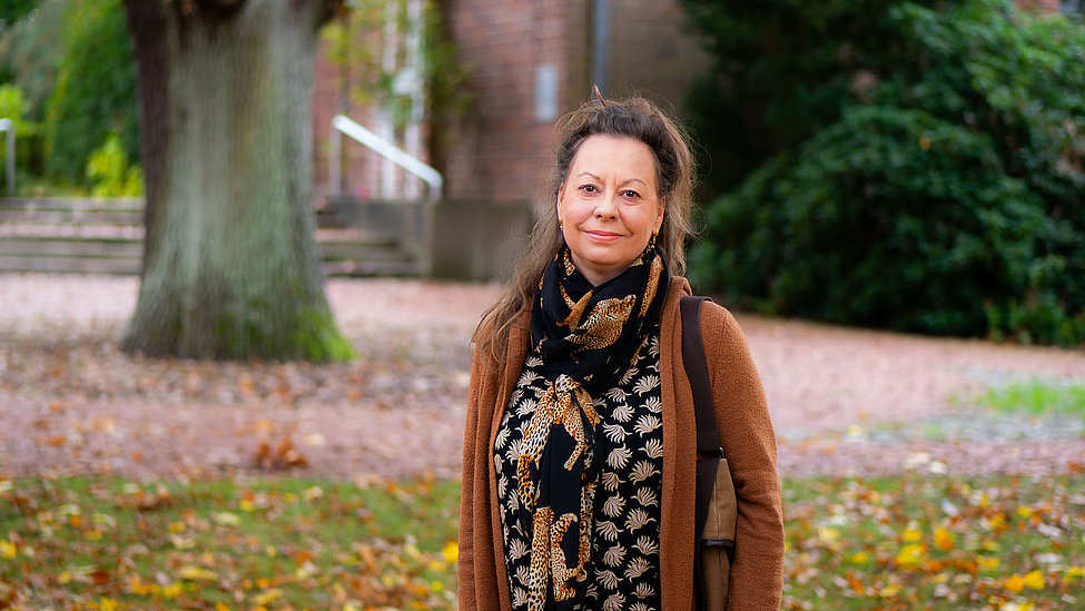 Auf dem Bild ist eine Frau zu sehen, die freundlich in die Kamera lächelt. Im Hintergrund sind in Unschärfe ein Gebäude, Büsche und Bäume zu erkennen. 